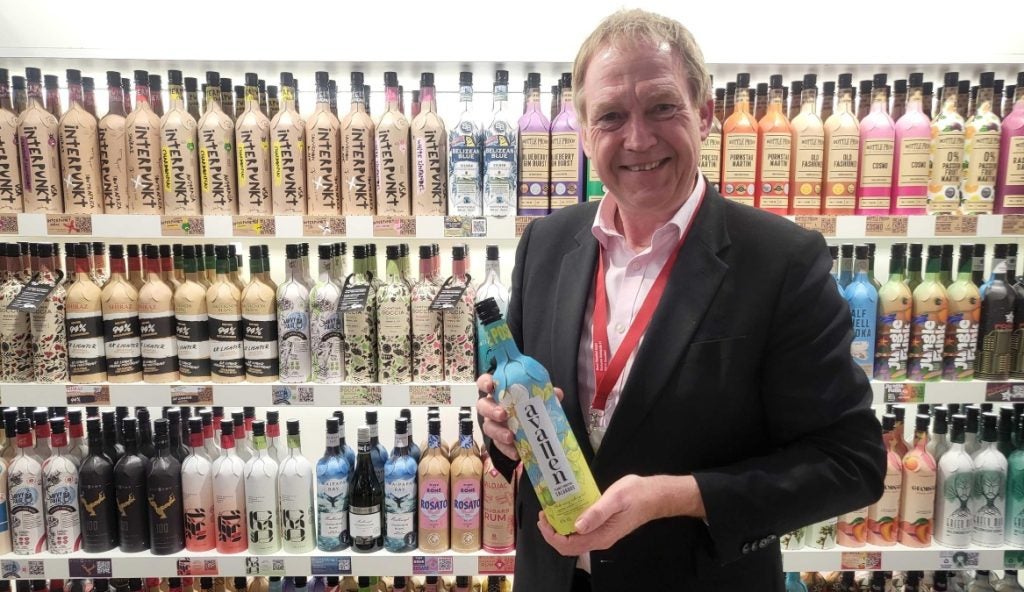 A man holds a paper Calvados bottle in front of shelves full of paper wine and spirits bottles