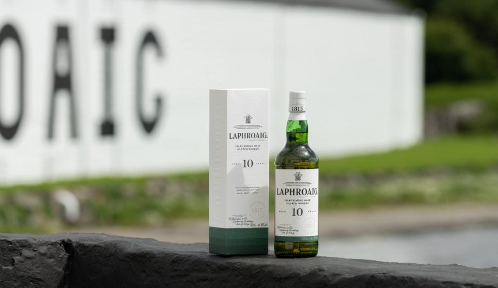 A bottle of Laphroaig Scotch whisky stands next to a gift box on a wall with the white distillery wall behind