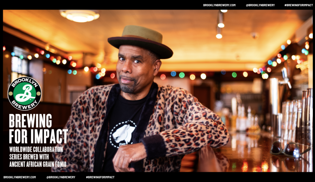 A black man in a leopard print jumper and boater hat smiles at the camera with his arm on a bar